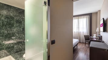 A hotel room with a glass shower enclosure, a bed, a desk, a chair, and a window with curtains.