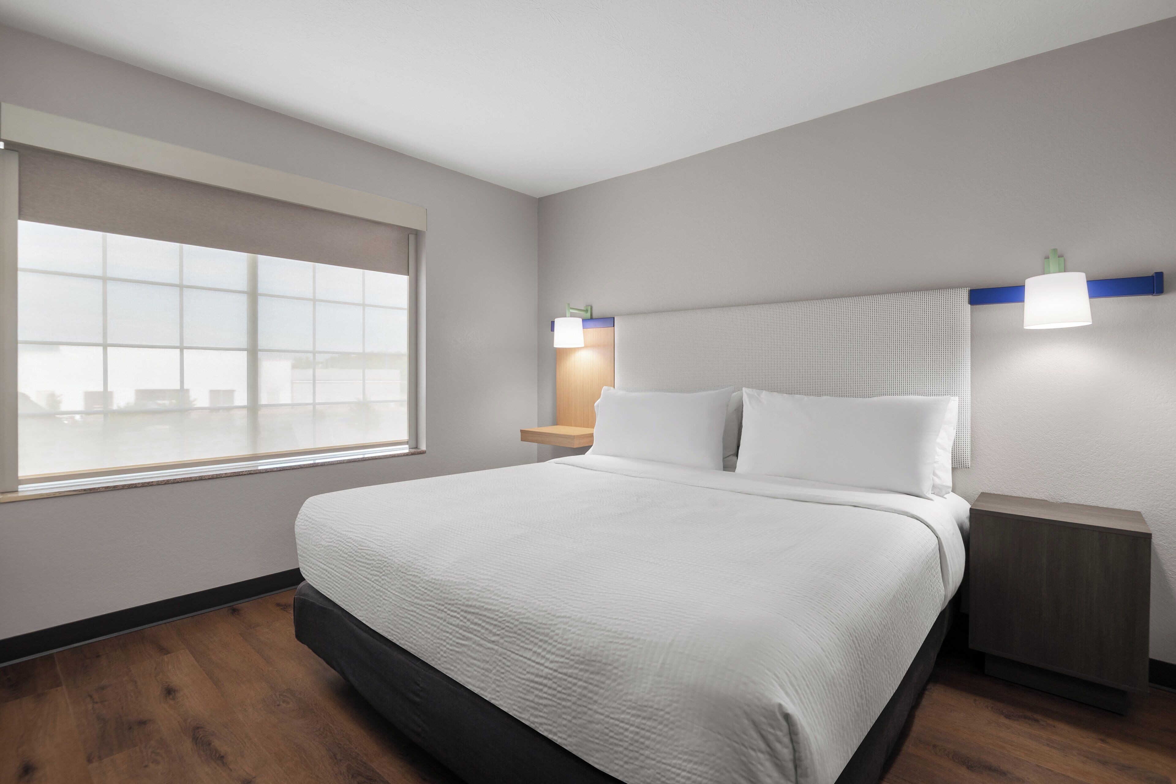 A modern hotel room with a large bed, a nightstand, a wall-mounted lamp, and a window with blinds.