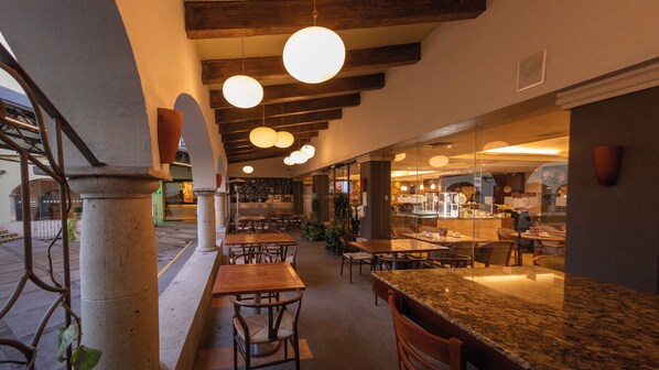 Un restaurante interior con mesas y sillas de madera, lámparas colgantes y una zona de bar con mostrador de granito.