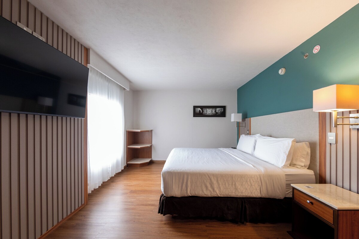 Una habitación de hotel con cama, mesita de noche, lámpara y un cuadro colgado en la pared.