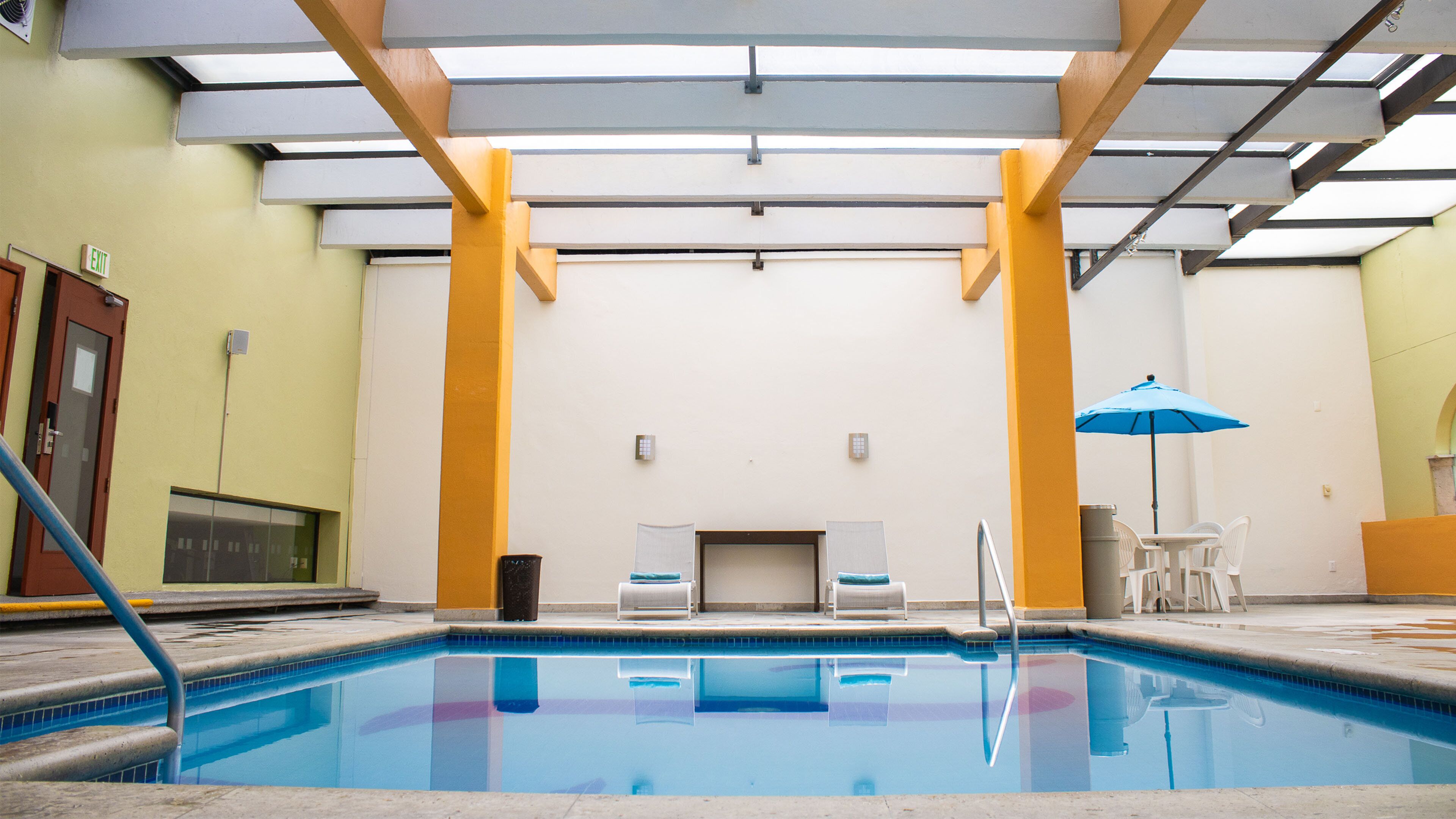 Una piscina con agua cristalina azul, rodeada de un interior blanco y amarillo, y una sombrilla azul.