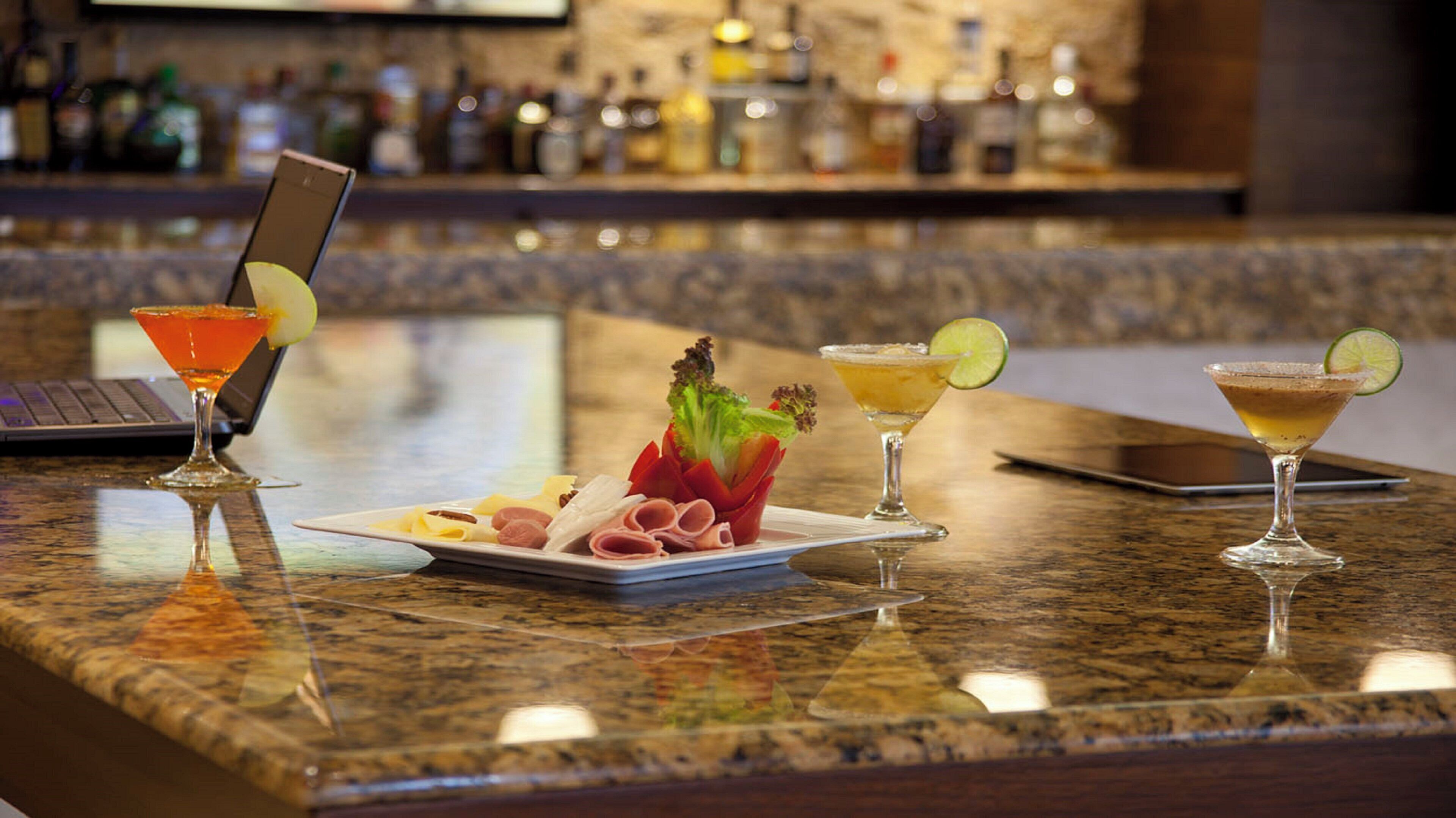 Um espaço de bar com um laptop, três coquetéis e um prato de comida.