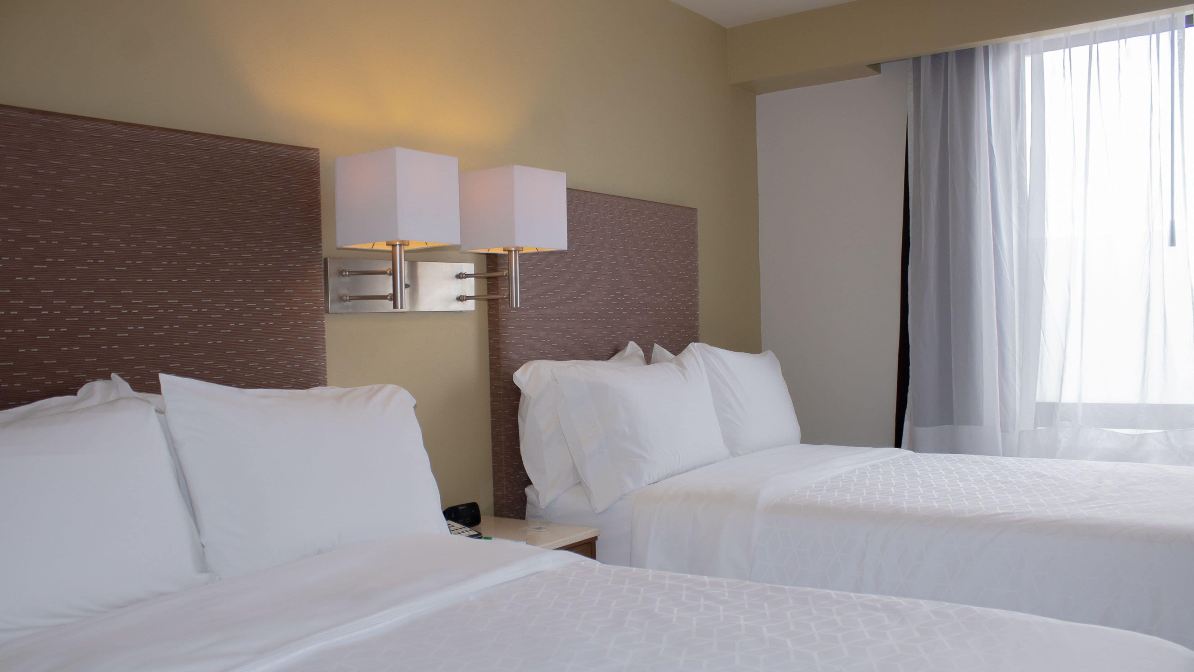 A hotel room with two beds, a wall-mounted light fixture, and a large window with curtains.
