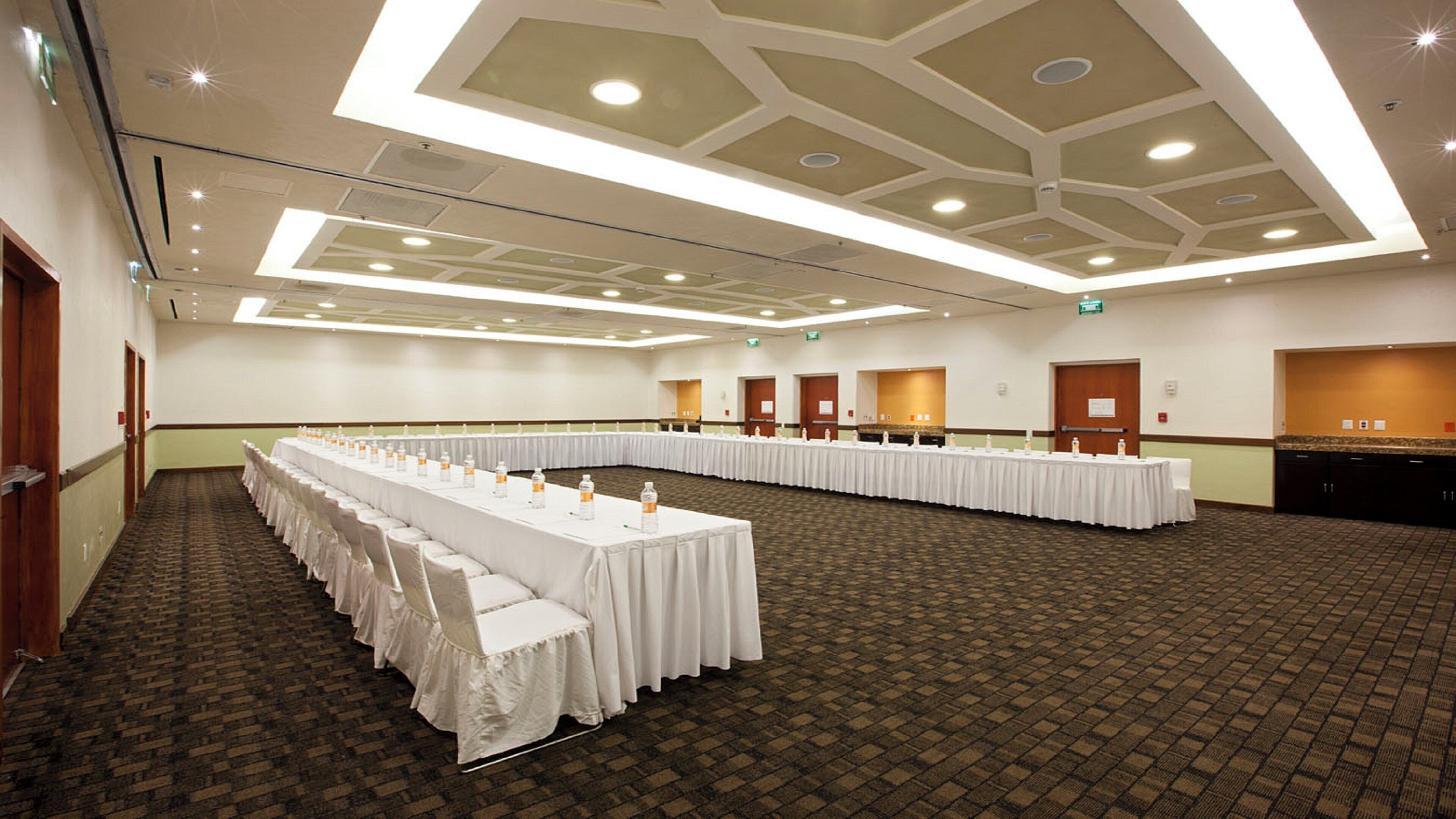 Un salón de conferencias con largas mesas, manteles blancos y botellas de agua.