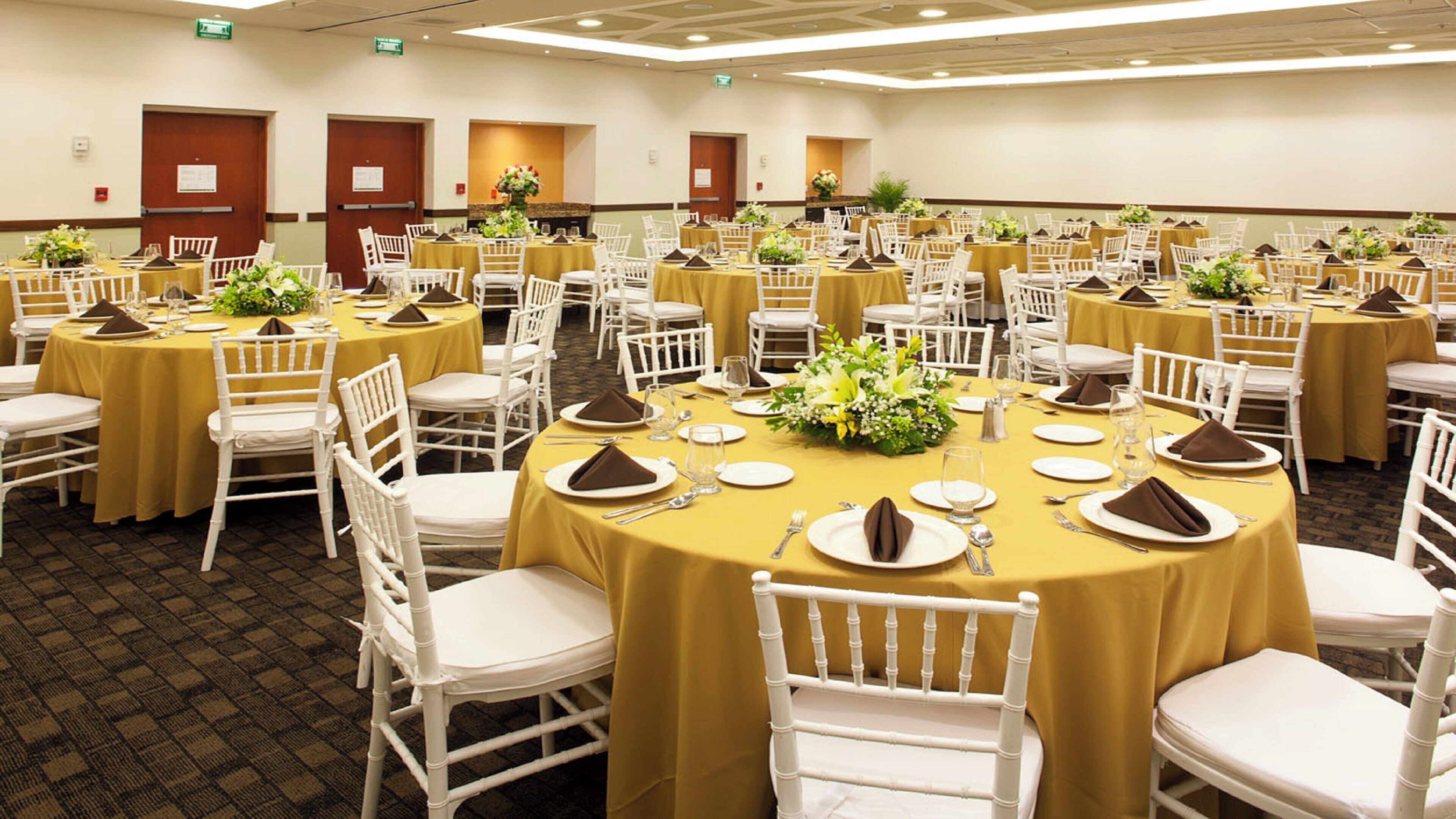 Un salón de conferencias con varias mesas redondas, cada una con sillas blancas y manteles amarillos.