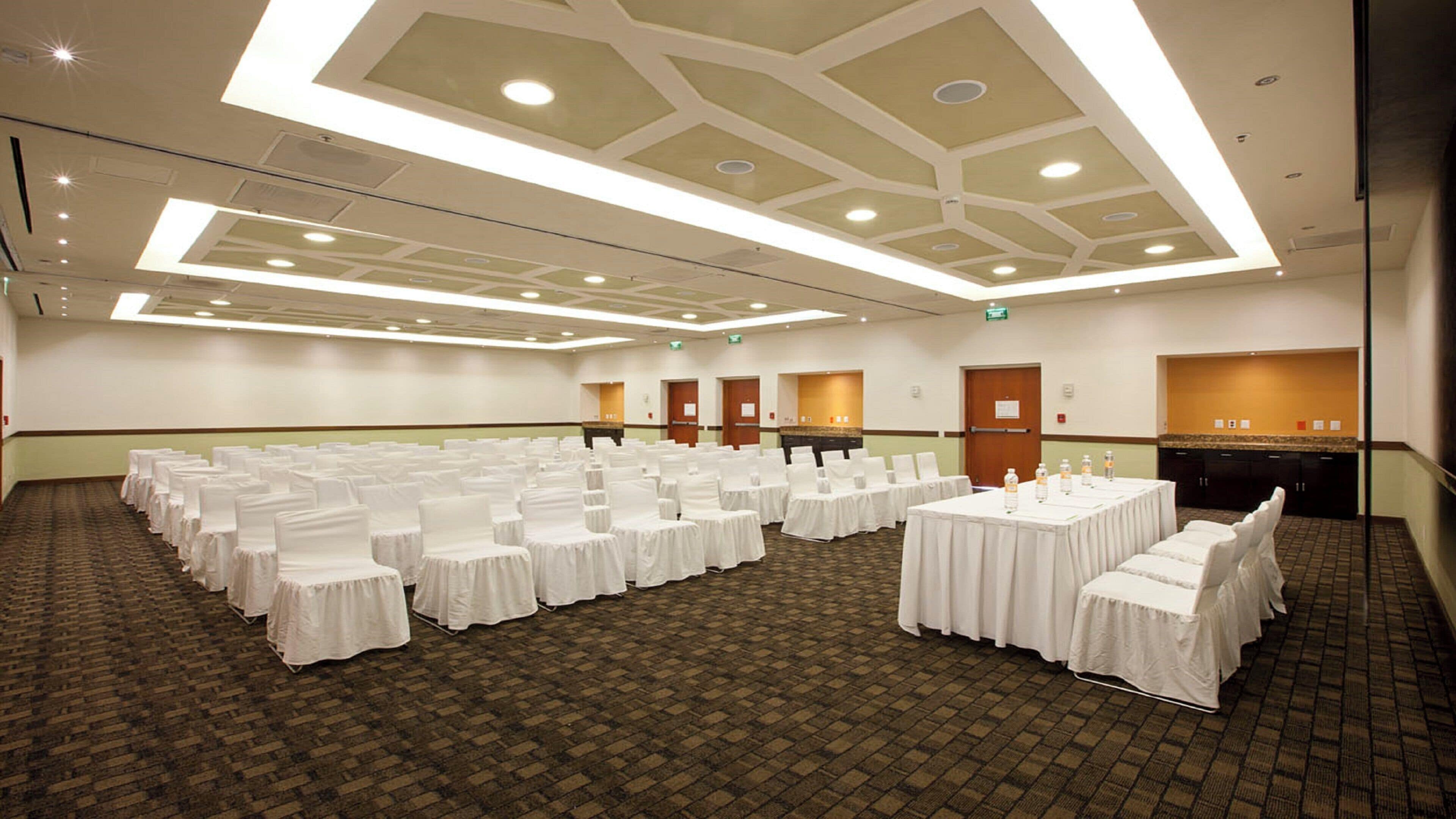 A conference room with rows of tables and chairs, a kitchenette, and a patterned carpet.
