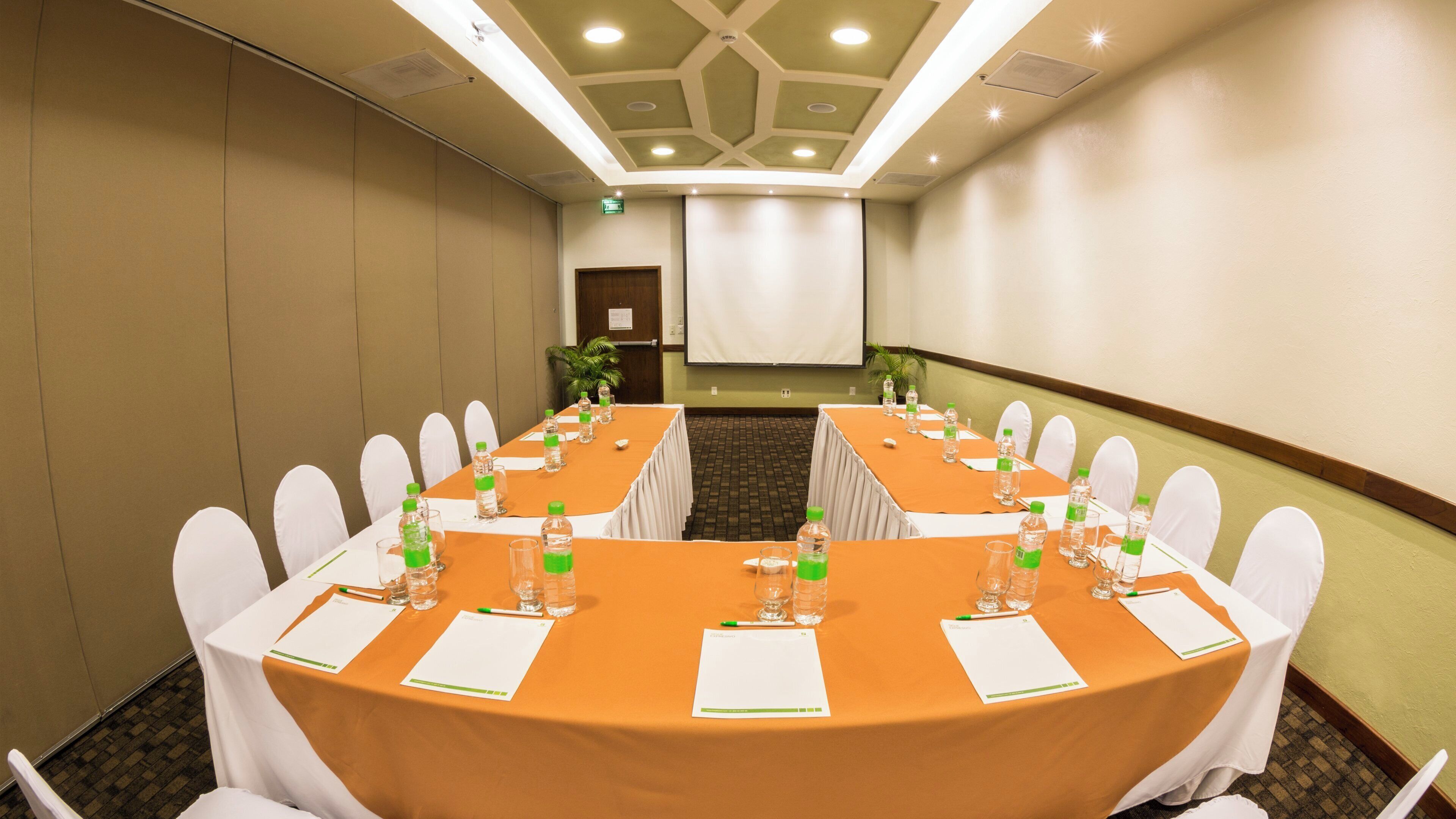 Un salón de conferencias con una larga mesa, sillas blancas y botellas de agua.