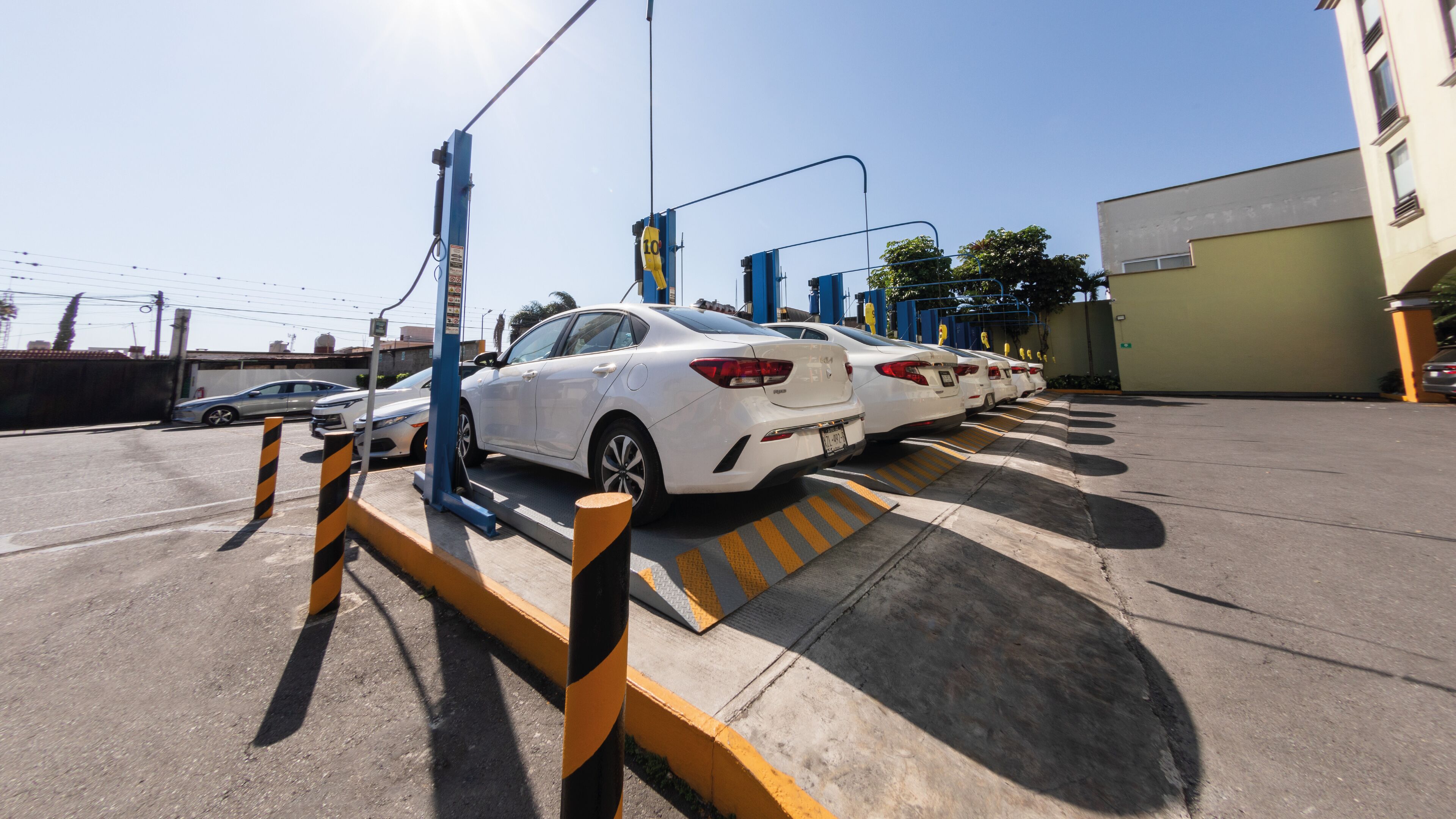 A parking area with multiple white cars parked in a row, a blue barrier gate, and a yellow and black striped safety bollard.