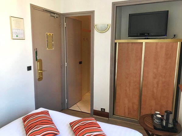 Habitación de hotel con una cama, un televisor sobre un mueble, una puerta con cerradura y una mesita con cafetera y tazas.