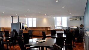 A modern dining area with round tables and chairs, a kitchen in the background, and large windows allowing natural light. - Hotel Colbert (Saint-Jean-de-Luz)