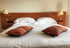 Una cama bien hecha con sábanas blancas y dos almohadas decorativas, un cabecero de madera y lámparas de noche.