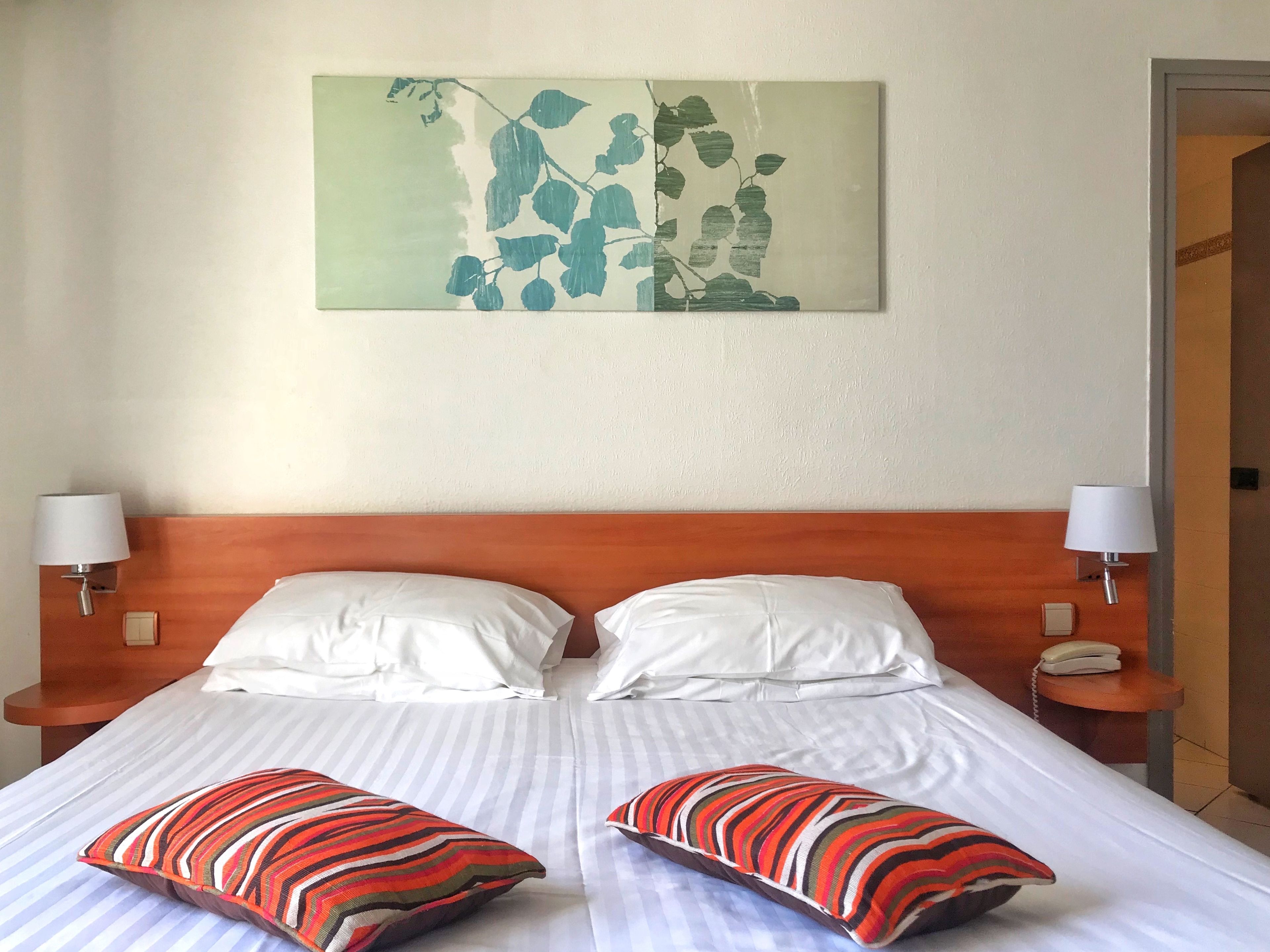 Ein Hotelzimmer mit einem hölzernen Kopfteil, zwei weißen Kissen, gestreiften Kissen, einem Bett mit weißer Bettwäsche, einem an der Wand befestigten Kunstwerk mit Blättern und Nachttischlampen.