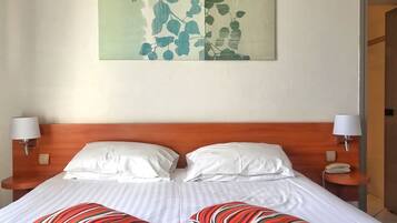 Ein Hotelzimmer mit einem hölzernen Kopfteil, zwei weißen Kissen, gestreiften Kissen, einem Bett mit weißer Bettwäsche, einem an der Wand befestigten Kunstwerk mit Blättern und Nachttischlampen.