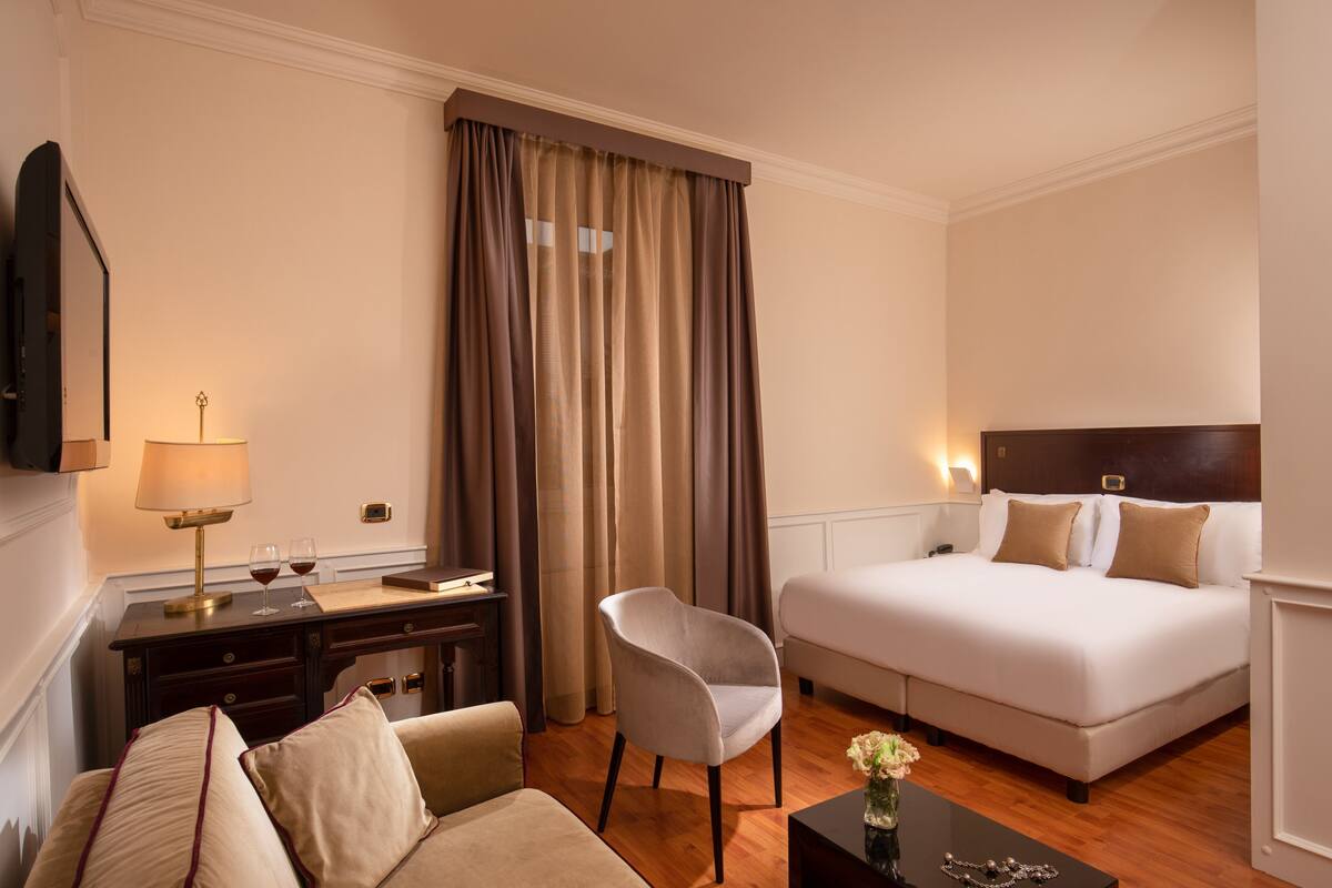 A hotel room with a bed, a desk with a lamp, a chair, and a small table with a vase.
