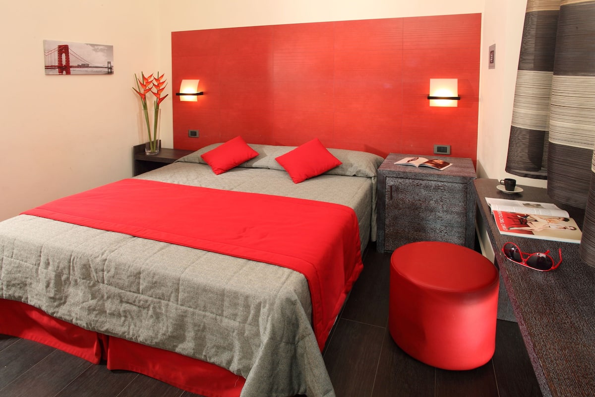 A hotel room with a large bed, two bedside tables, a red ottoman, and a wall-mounted shelf with a magazine and sunglasses.