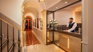 Un hall d’hôtel avec un escalier en marbre, un comptoir d’accueil et un couloir menant à une pièce dotée d’une grande porte en arc de cercle.
