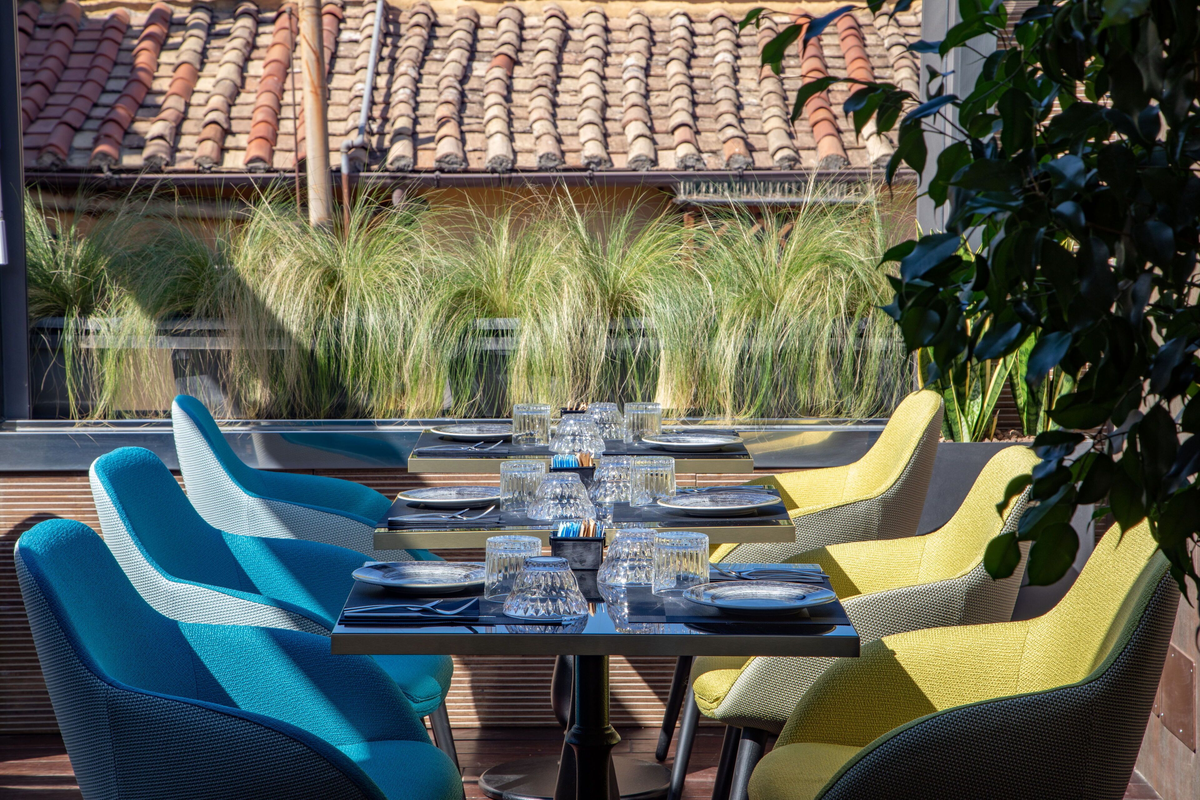 An outdoor dining area with a table set for six, surrounded by blue and yellow chairs, overlooking a garden with tall grasses and a tiled roof in the background.