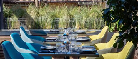 An outdoor dining area with a table set for six, surrounded by blue and yellow chairs, overlooking a garden with tall grasses and a tiled roof in the background.