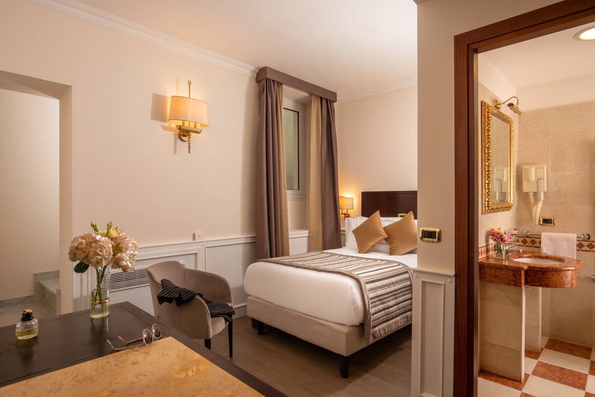 A hotel room with a bed, bedside table, chair, and a bathroom with a sink and mirror.