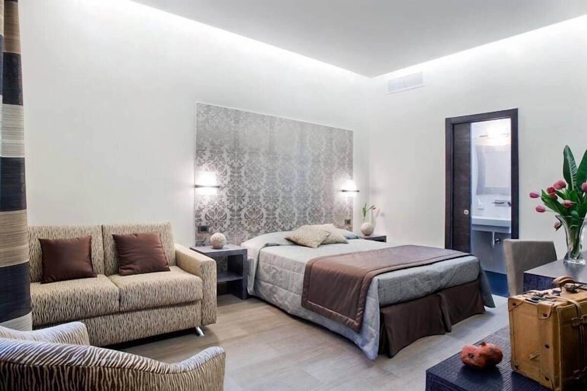 A modern hotel room with a large bed, a sofa, a desk, and a bathroom visible through an open door.