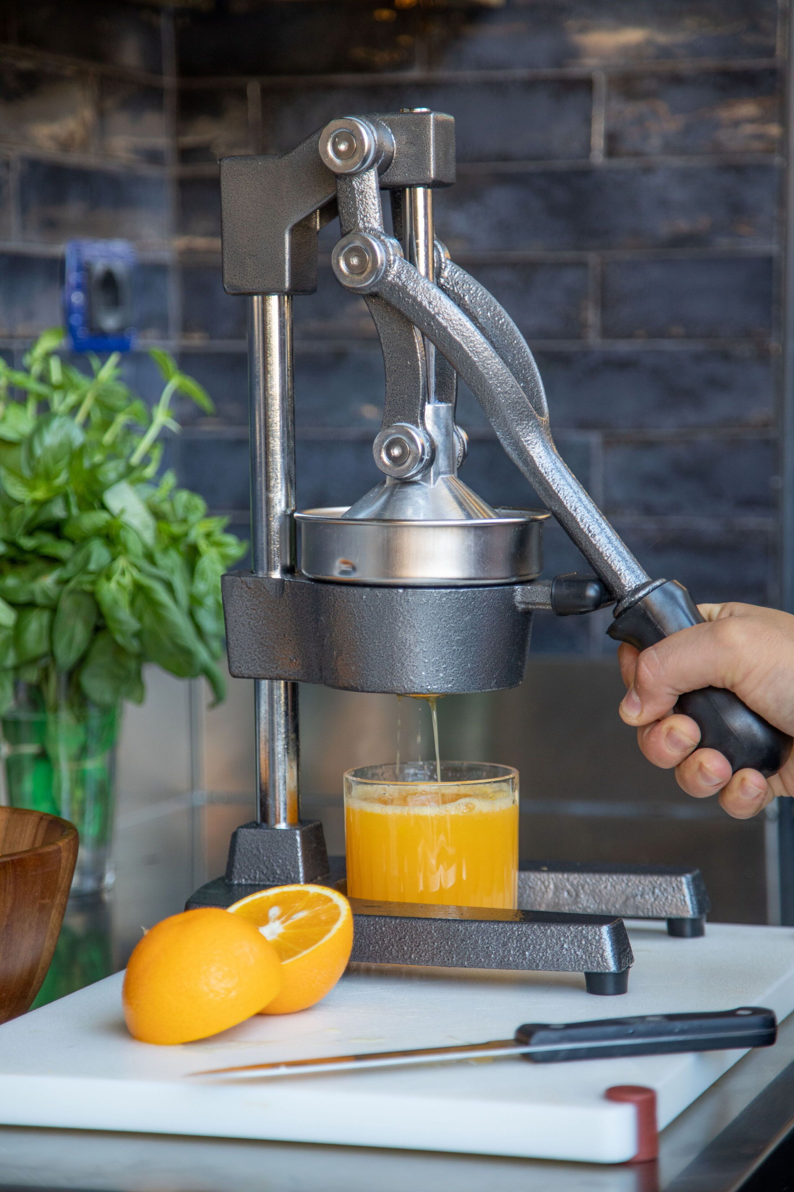 A manual juicer with a hand squeezing it, a glass of juice, and two oranges on a cutting board.
