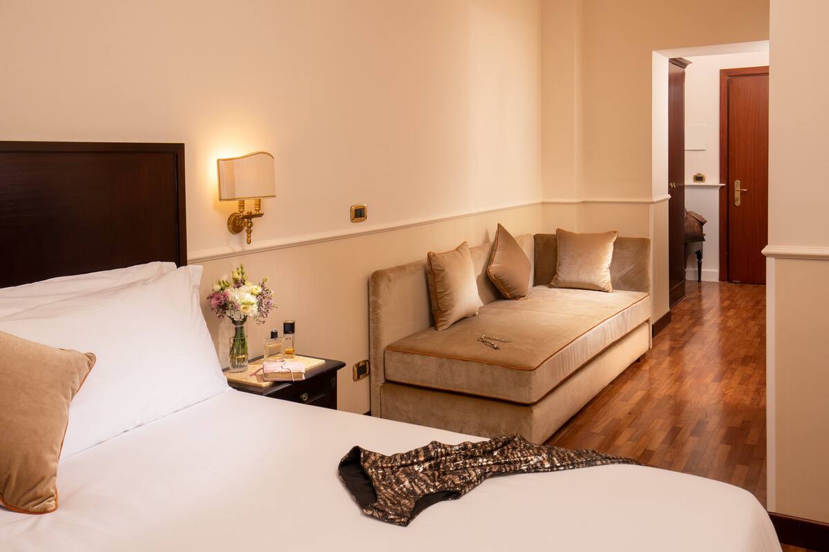 A hotel room with a bed, a sofa, a nightstand with a lamp, and a vase of flowers.