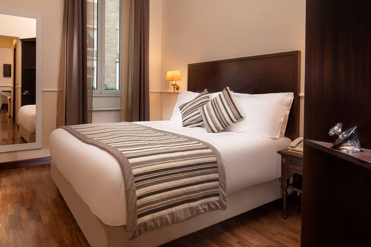 A hotel room with a large bed, a bedside lamp, a mirror, and a wooden floor.