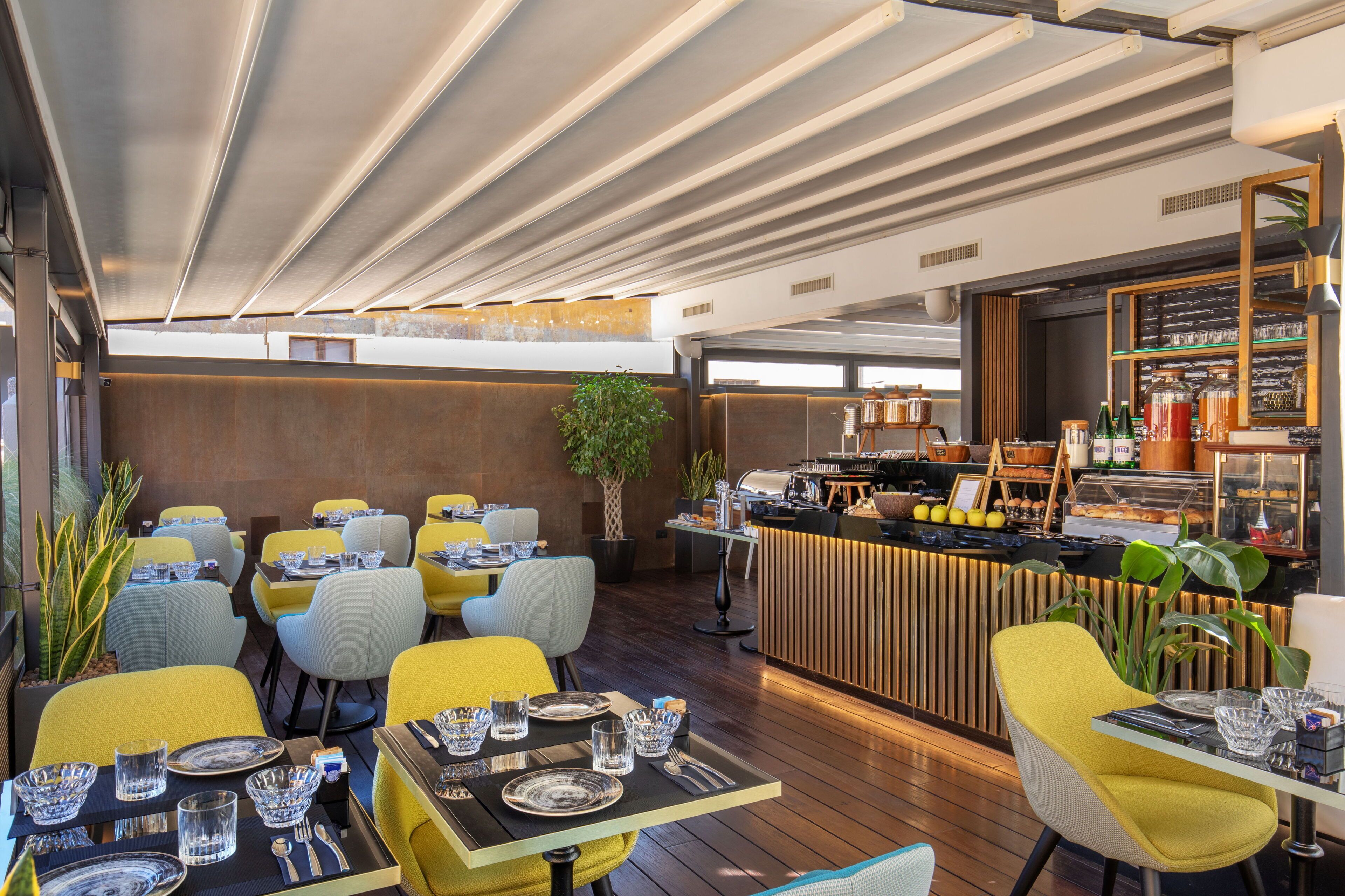 Un restaurante moderno con sillas amarillas y grises, mesas de madera y una zona de bar central con una exhibición de alimentos.