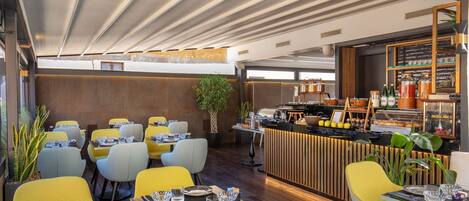 Un restaurante moderno con sillas amarillas y grises, mesas de madera y una zona de bar central con una exhibición de alimentos.