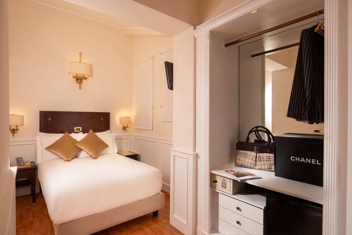 A hotel room with a neatly made bed, bedside lamps, a dresser with a Chanel bag, and a mirror reflecting a striped garment.