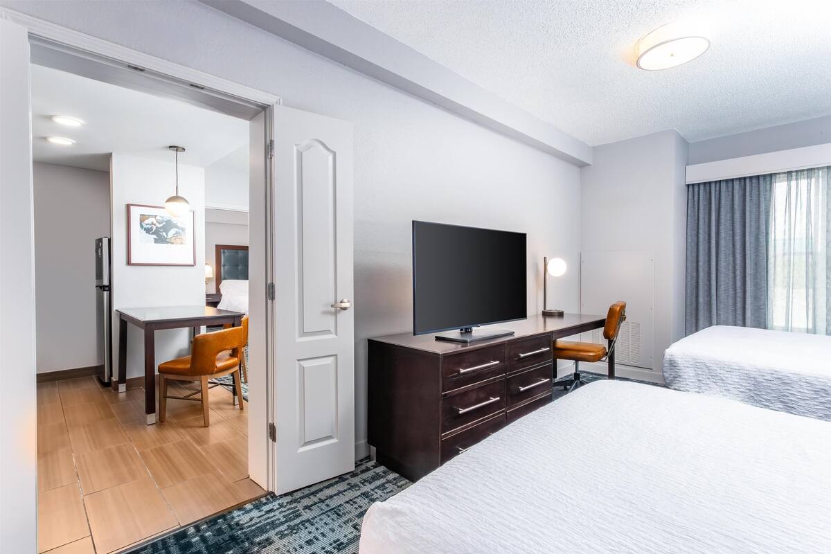Een hotelkamer met een bed, een televisie op een dressoir, een bureau met stoel, en een deur die naar een andere kamer leidt.