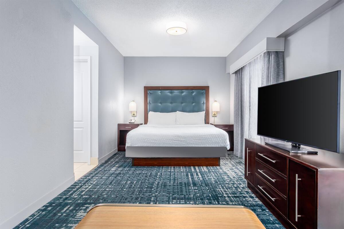Hotelkamer met een groot bed, een flatscreen televisie, een houten bankje en een kamerkleed met een patroon.