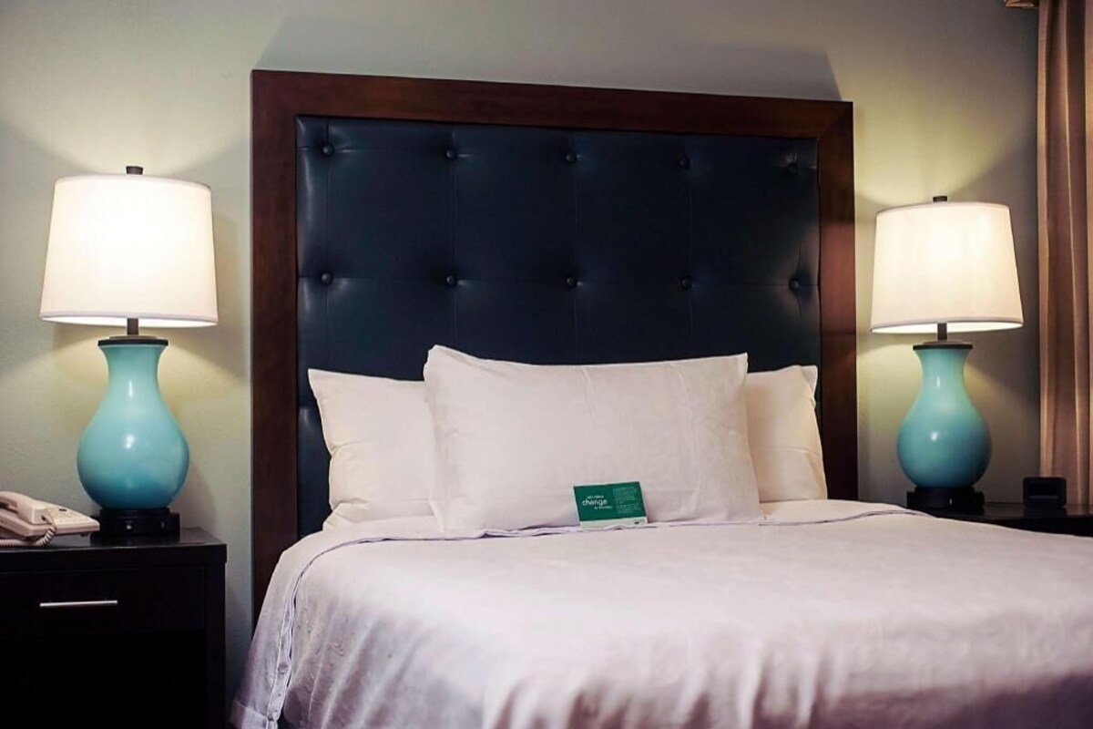 A hotel room with a tufted headboard, two bedside lamps, a bed with white linens, a bedside phone, and a green tag on the bed.