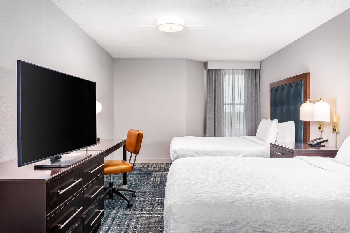 Een hotelkamer met een flatscreen televisie, een bureau met lades, twee bedden, een stoel, een lamp en een raam met gordijnen.