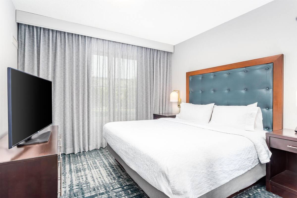 Een hotelkamer met een groot bed, een flatscreen televisie, een nachtkastje, een lamp en een raam met gordijnen.