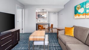 Una habitación de hotel moderna con televisor de pantalla plana, una mesa de comedor de madera, un sofá con un cojín amarillo y una cocina al fondo.