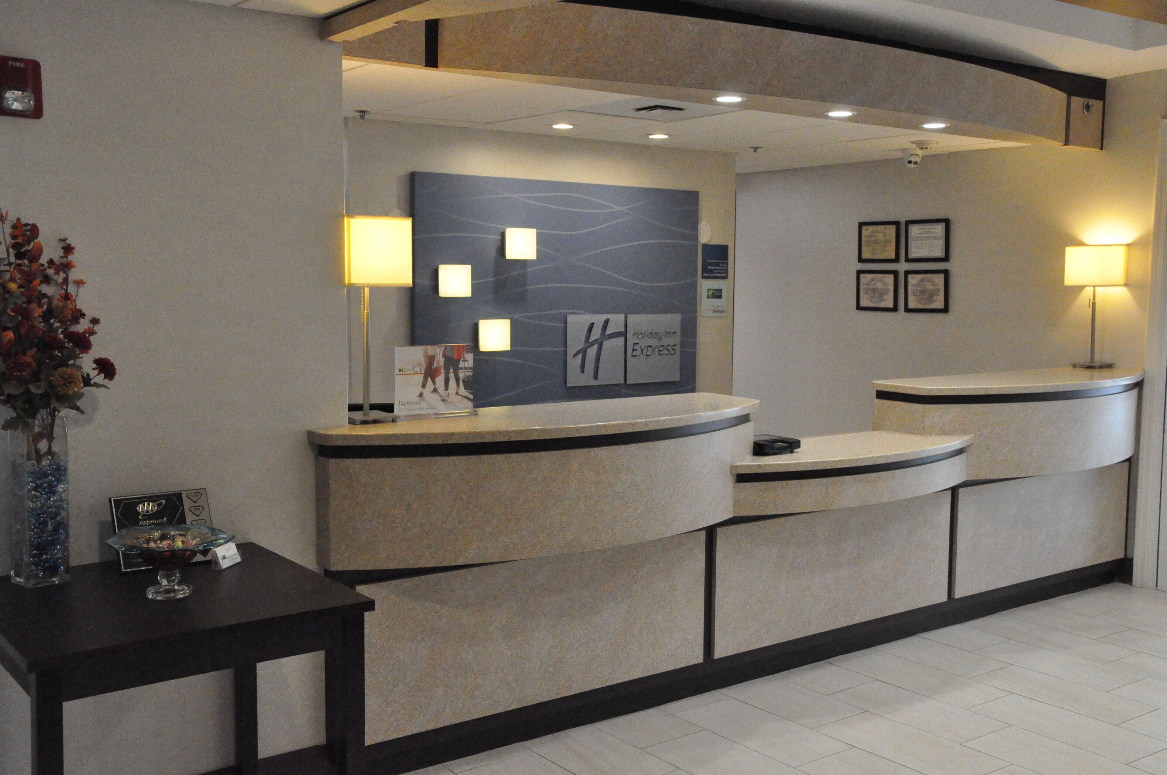 Un mostrador de recepción de hotel con diseño moderno, que presenta una distribución curva y un logotipo central. Hay dos lámparas fijadas a la pared que proporcionan iluminación.
