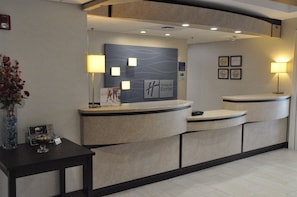 A hotel reception desk with a modern design, featuring a curved layout and a central logo. There are two wall-mounted lamps providing illumination. - Holiday Inn Express Hotel & Suites Jackson by IHG (Jackson)