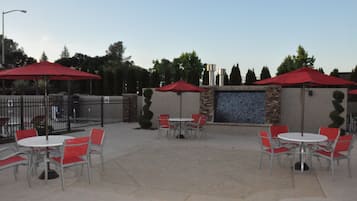 Un área de patio al aire libre con mesas y sillas, sombrillas rojas y una pared decorativa.