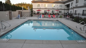 A rectangular swimming pool with a tiled deck, surrounded by a metal fence, and a building with multiple windows and air conditioning units in the background.