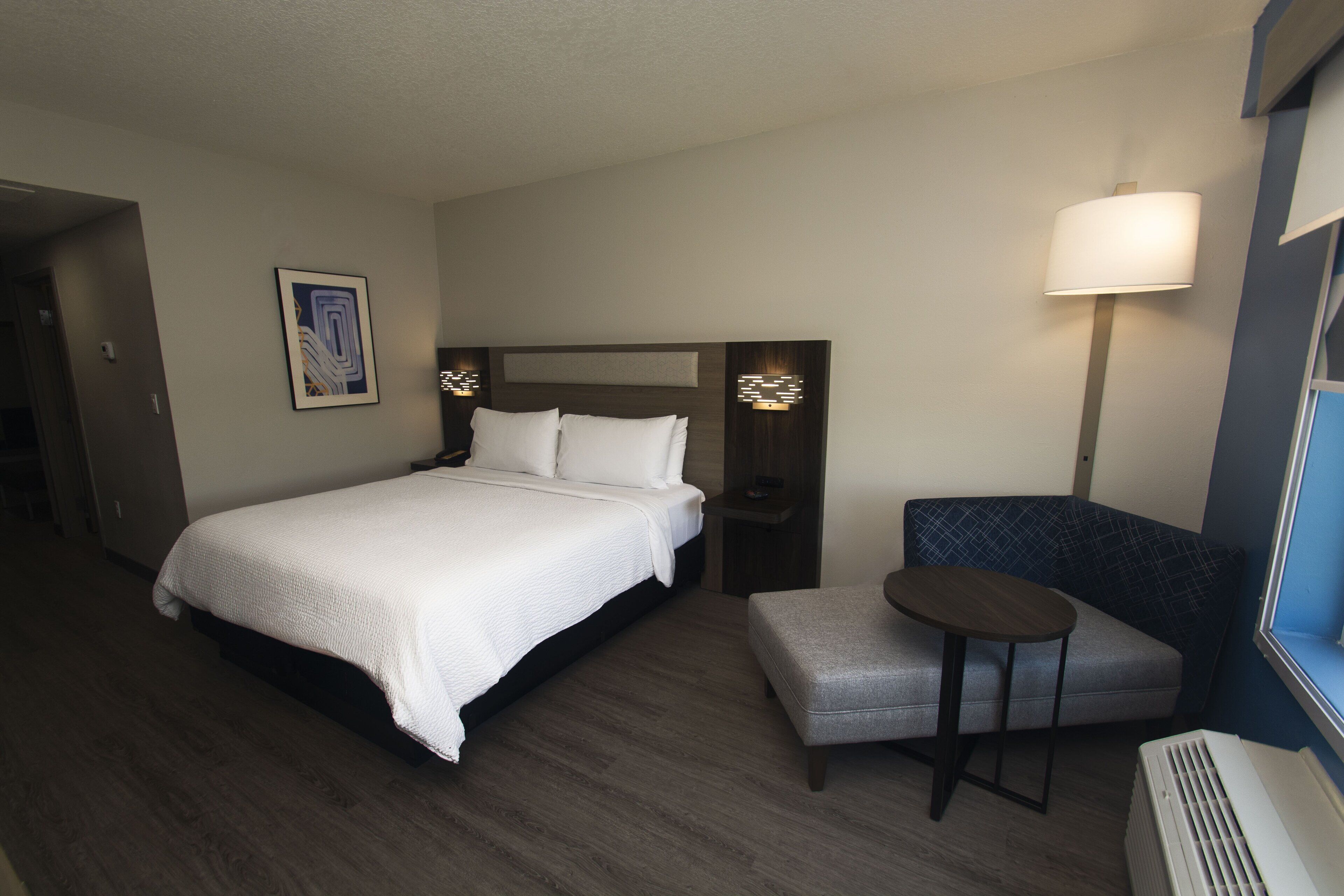 A hotel room with a bed, a small table, a chair, and a wall-mounted lamp.