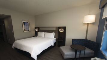 A hotel room with a bed, a small table, a chair, and a wall-mounted lamp.