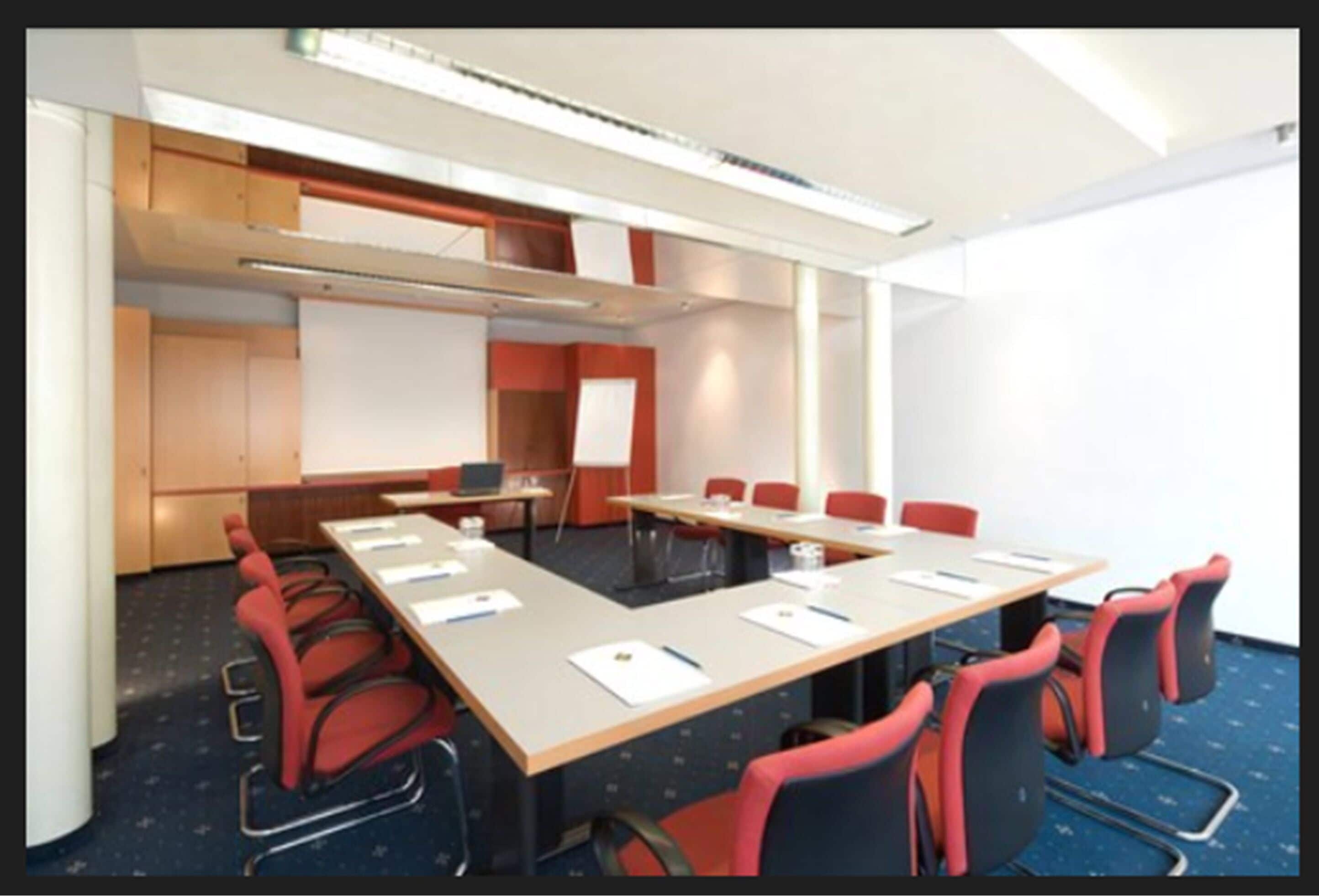 A conference room with a long table, red chairs, a whiteboard, and a projector screen.