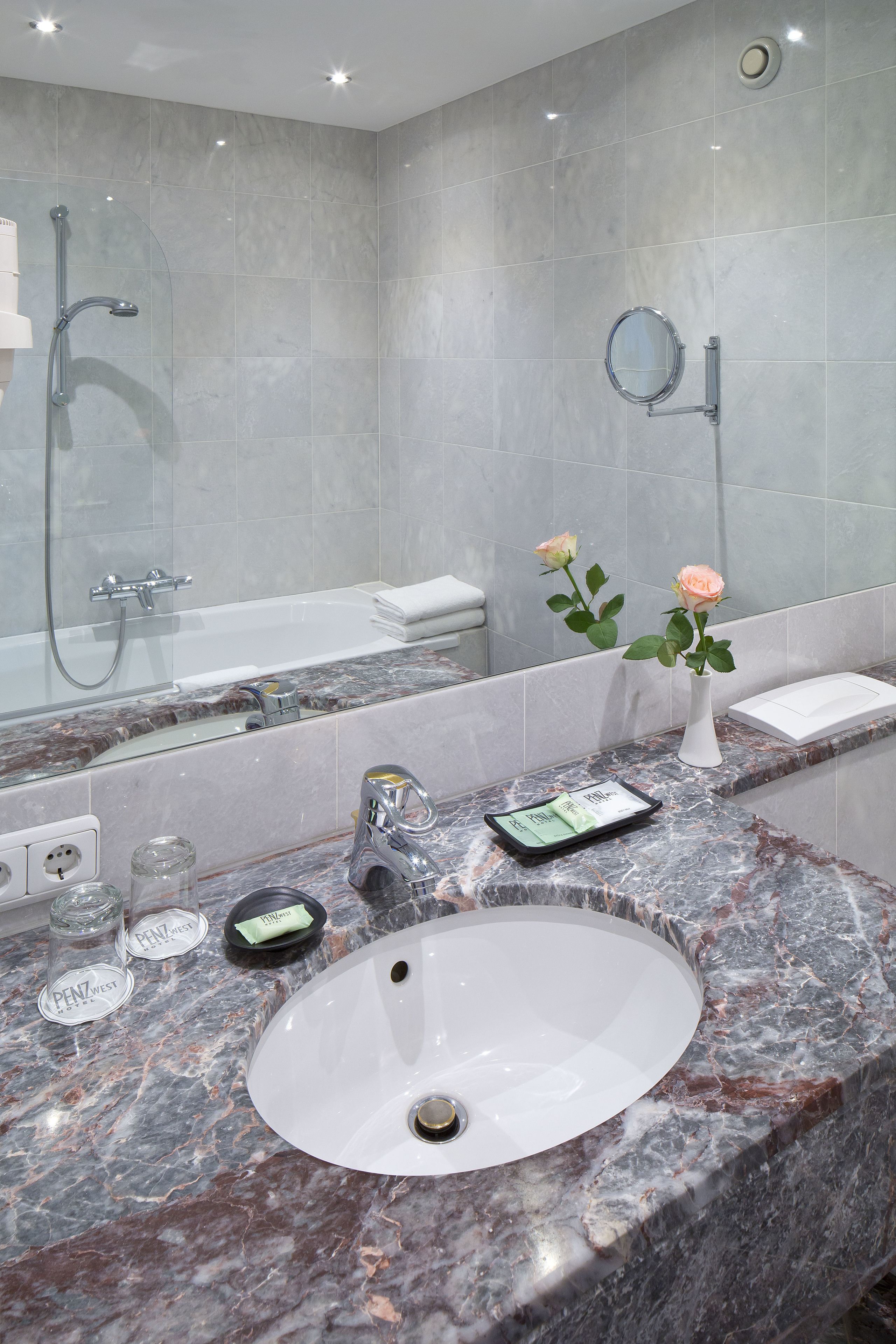 Un baño con lavamanos de mármol, cabezal de ducha de mano, espejo redondo y un florero con flores.