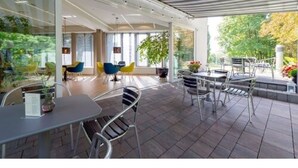 Un patio exterior con mesas y sillas, amplias ventanas y plantas en macetas.