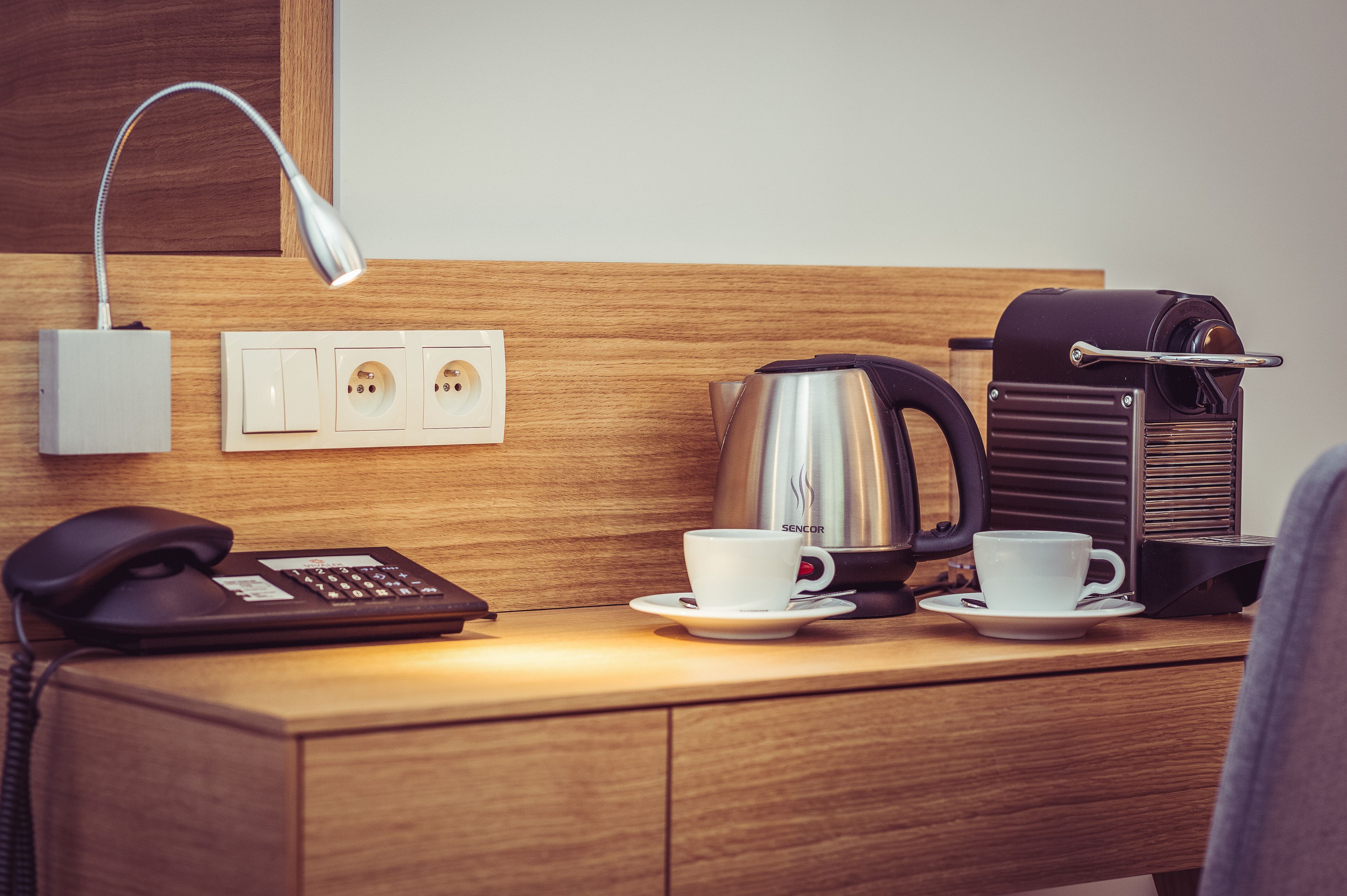 Et natbord i træ med en lampe, kaffemaskine, kop, underkop og en telefon.