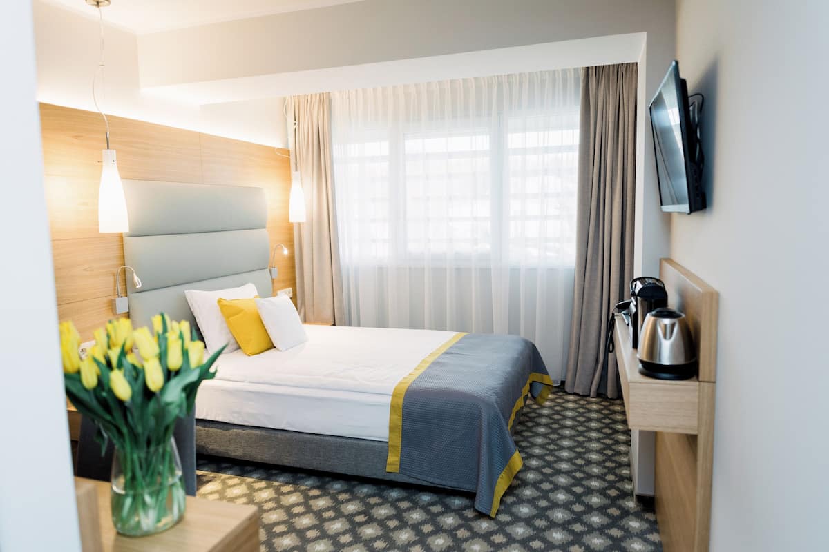 Una habitación de hotel con una cama, una mesita de noche con un hervidor, un televisor montado en la pared y un jarrón con flores amarillas.