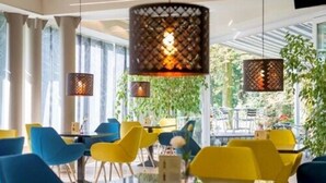 Un comedor moderno con sillas amarillas y azules, lámparas colgantes y grandes ventanas que dan a vistas de follaje.