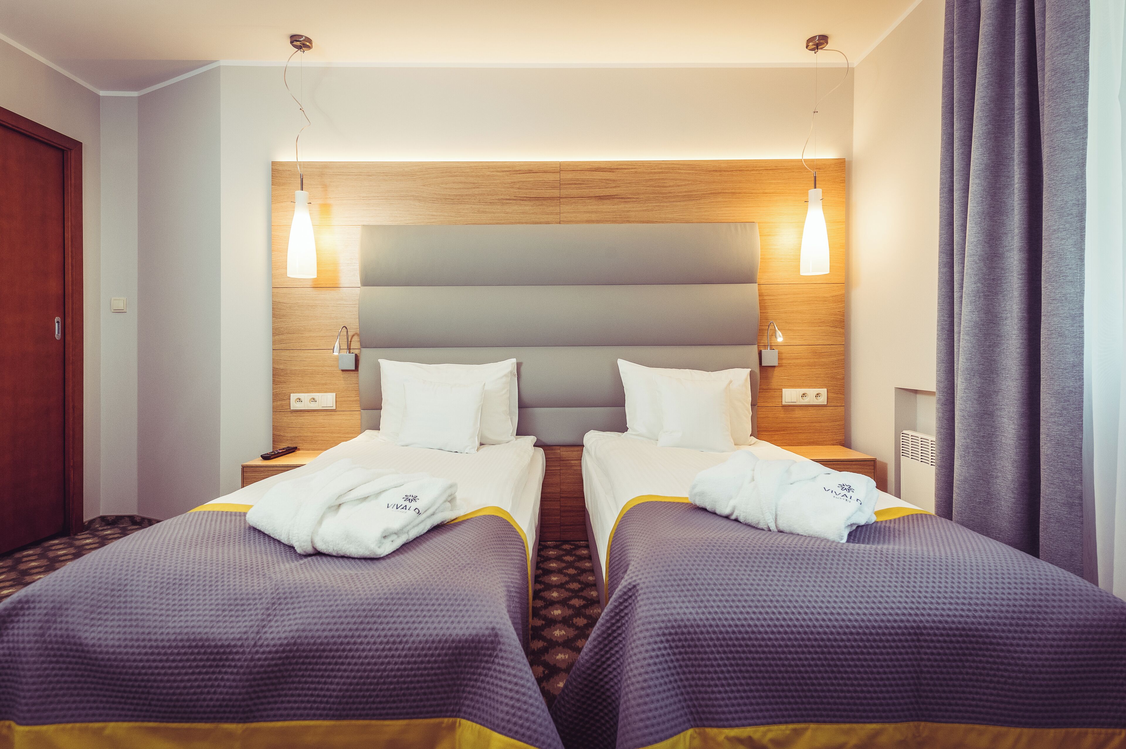 Habitació d'hotel amb dos llits, un panell de capçalera de fusta, làmpades laterals i una tauleta de nit amb un comandament a distància.