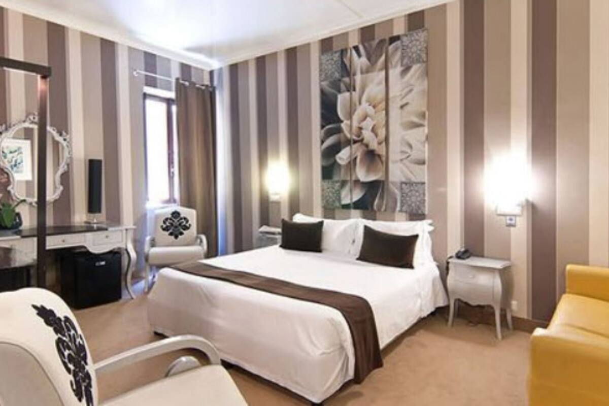 Ein Hotelzimmer mit einem großen Bett, einem Nachttisch, einem Sessel, einem Schreibtisch, einem Spiegel und einem großen Blumenbild an der Wand.